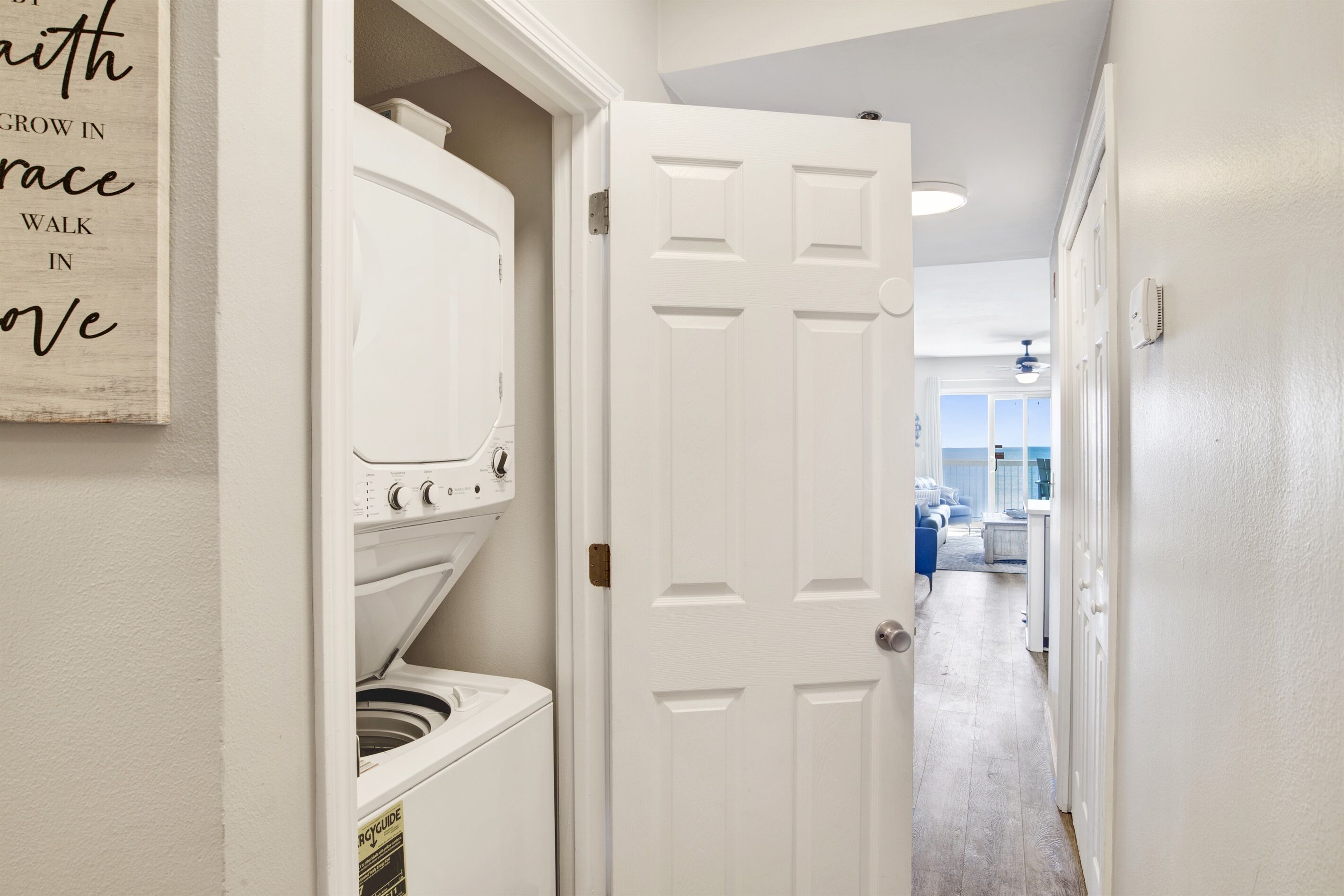 A laundry room with a front-loading washer and dryer, a white door, and a hallway leading to a living area.