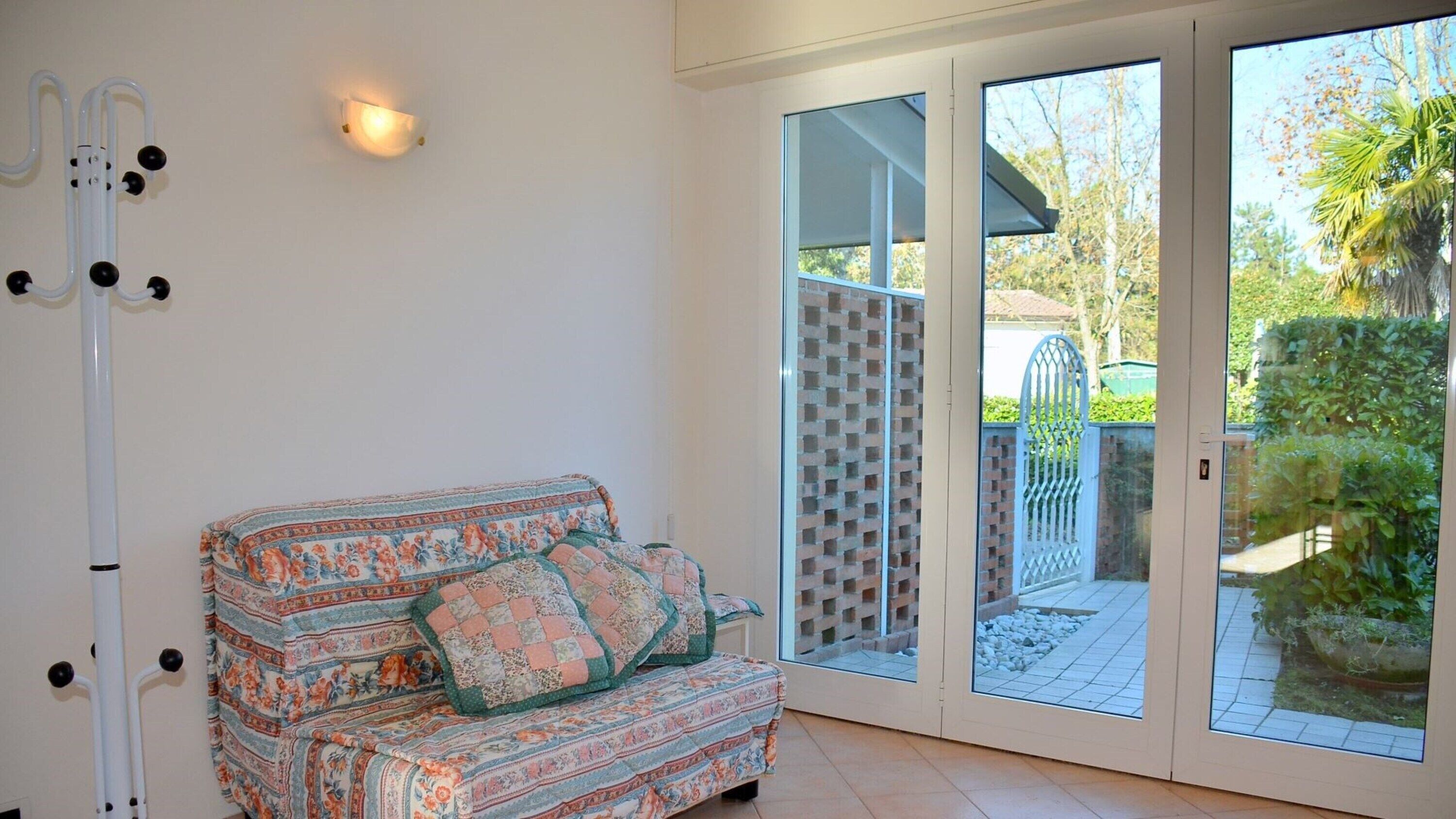 A room with a patterned sofa, a white wall, a ceiling-mounted light fixture, and a sliding glass door leading to an outdoor area with a brick wall and greenery.