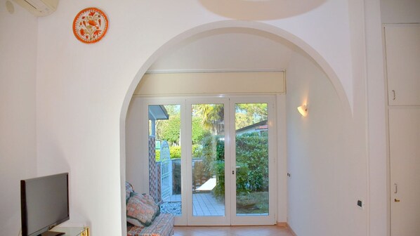 A room with a television, a patterned rug, a white door, and a glass door leading to an outdoor area with a patio and greenery.