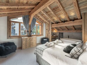 Ein gemütliches Schlafzimmer mit Holzbalkendecke, einem großen Fenster mit Vorhängen, einem Bett mit Kissen und einem kleinen Holztisch.
