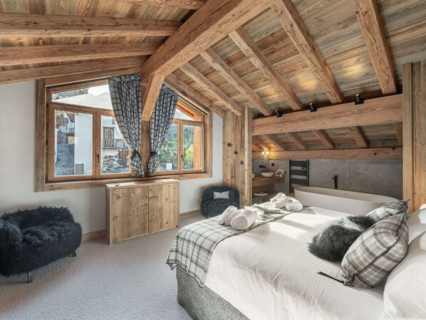 A cozy bedroom with a wooden ceiling, a large window with curtains, a bed with pillows, and a small wooden table.