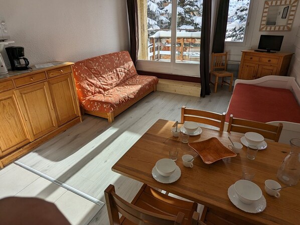 Une pièce chaleureuse avec une table à manger en bois, préparée pour quatre personnes, un canapé et un téléviseur posé sur un meuble en bois.