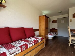 Un cuarto acogedor con un sofá cama de madera cubierto con una manta de diseño rojo y blanco, un armario de madera, una pequeña mesa de madera y una puerta que da a otra habitación.