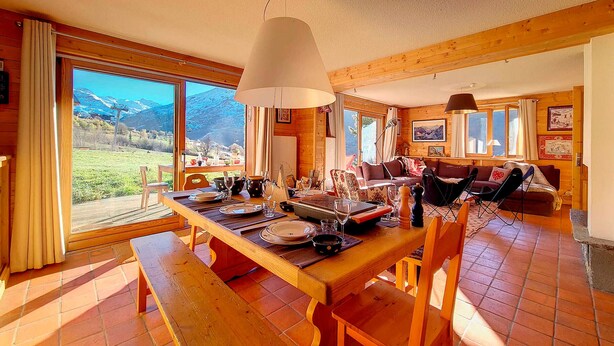 Ein Holzinterieur mit Esstisch für vier Personen, einer Couch und einem großen Fenster mit Blick auf die Berge.
