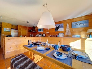 Interior de uma cabana de madeira com área de jantar posta para uma refeição, apresentando uma mesa com pratos azuis e brancos, um lustre pendente e paredes de madeira.