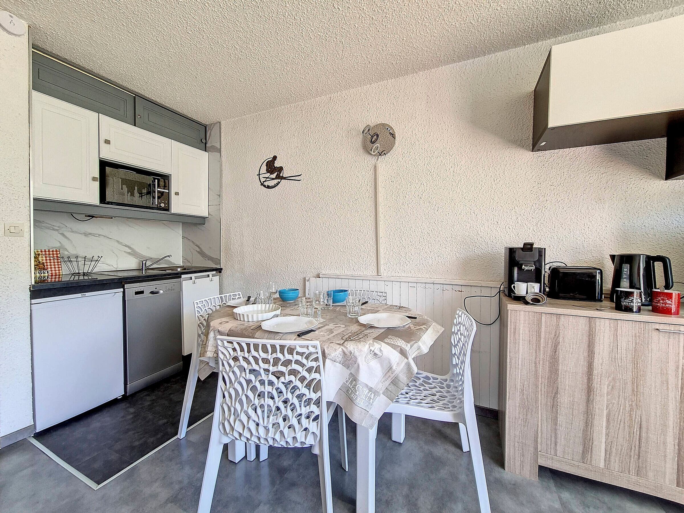 Une cuisine compacte avec des meubles blancs, un four à micro-ondes et une machine à café. On y trouve une table à manger avec des chaises et une horloge murale décorative.