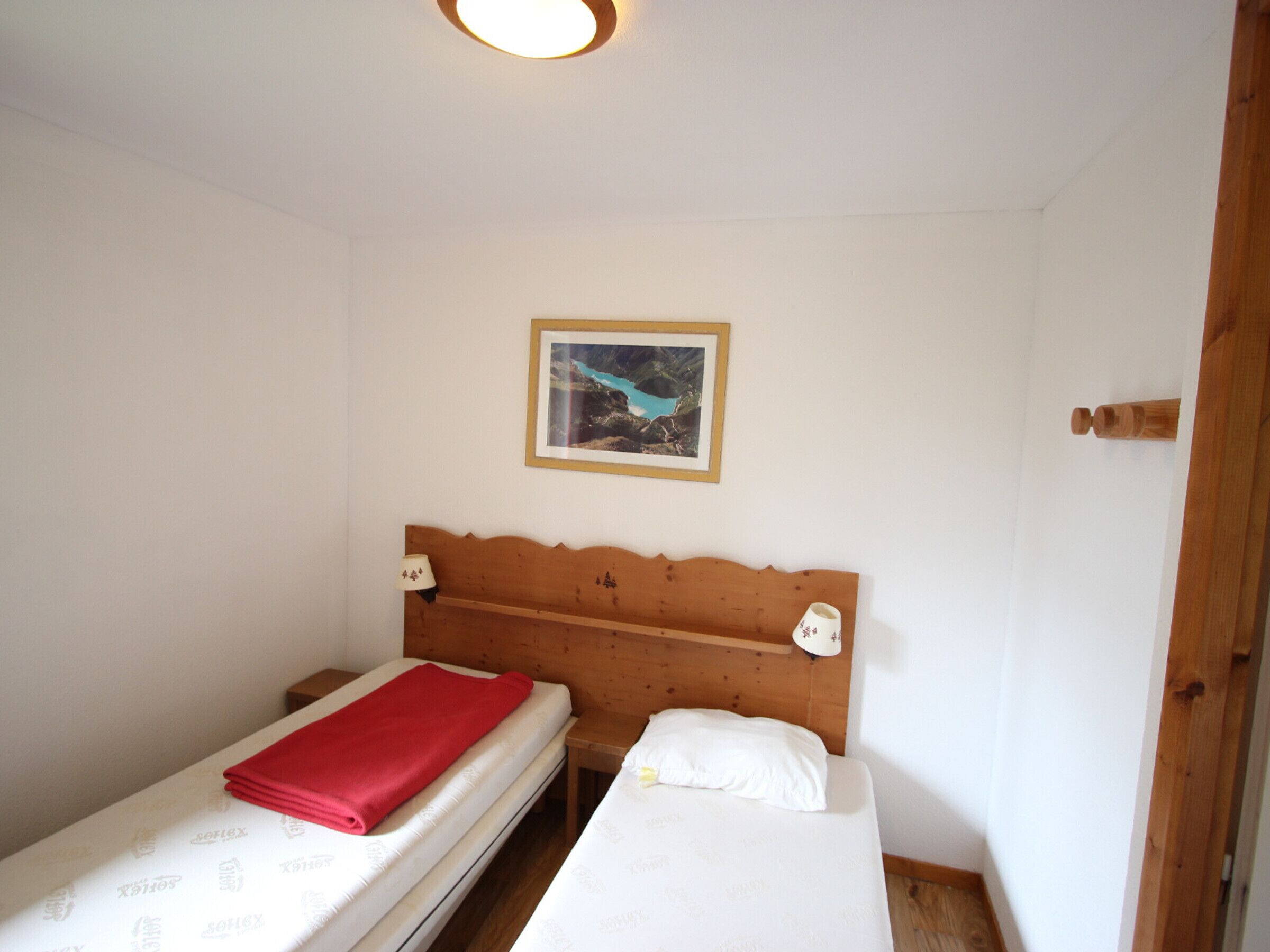 A small, simple room with two single beds, a wooden headboard, a framed picture on the wall, and a small table with a lamp.