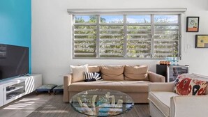 A living room with a beige sofa, a glass coffee table, a TV on a stand, and a window with blinds. - Pao de Azucar | Surf Haven with Spectacular Views (Rincón)