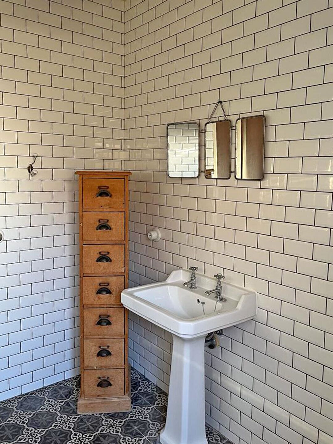 A bathroom with white subway tiles, a wooden chest of drawers, a white sink, and two mirrors.