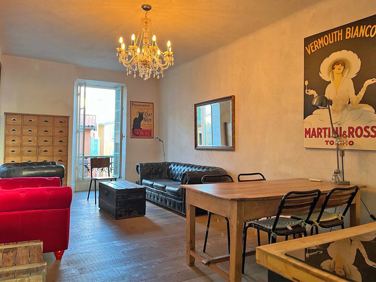 A living room with a red sofa, a black leather couch, a wooden dining table with chairs, a chandelier, and a poster on the wall.