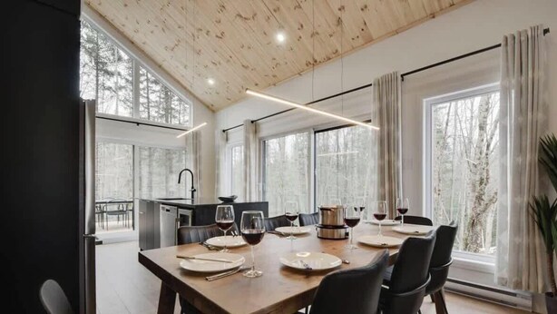 A dining area with a wooden table set for six, large windows, and a view of trees.