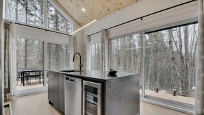 A modern kitchen with large windows, a central island, and a dining area with a view of a forest.