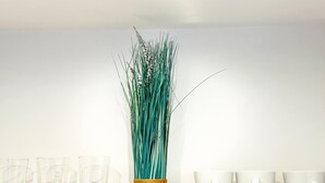 Uno scaffale con un vaso alto pieno di erba verde, diversi bicchieri trasparenti e tazze bianche.