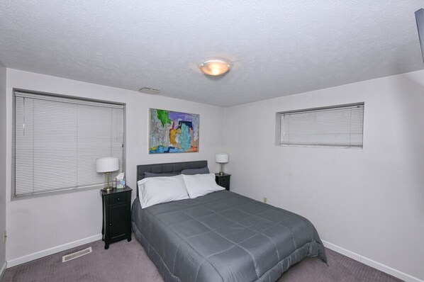 A bedroom with a bed, two bedside tables, a ceiling light, and a window with blinds.