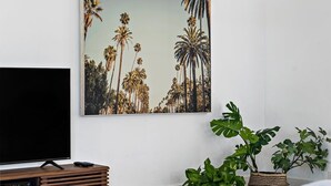 A modern living room with a flat-screen TV on a wooden stand, a large framed picture of palm trees on the wall, and two large green plants in woven pots. - Bohemian Condo Near Las Olas and Beach (Fort Lauderdale)