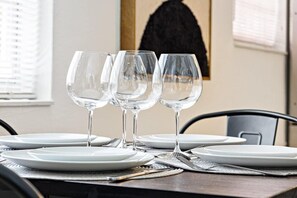 A dining table for four, complete with white plates and clear wine glasses, sits beside a black chair and a window with partially drawn blinds.