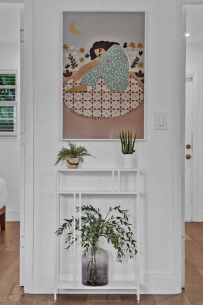 A white wall with a framed artwork of a woman sleeping, a small potted plant, and a shelf with a larger plant.