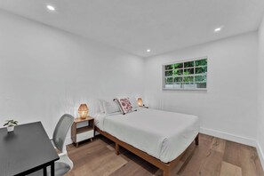 Une chambre à coucher avec un lit, des lampes de chevet, un bureau et une fenêtre donnant sur la verdure.
