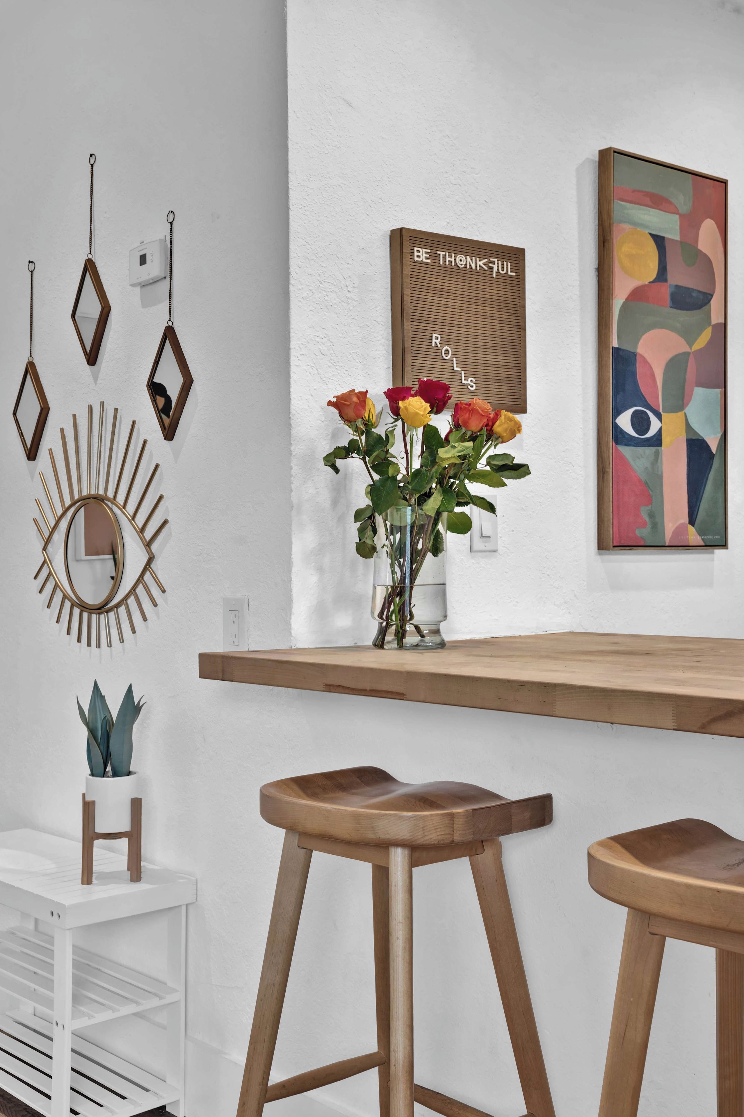 A modern interior with a wooden bar, bar stools, a mirror, a vase with flowers, and wall art.