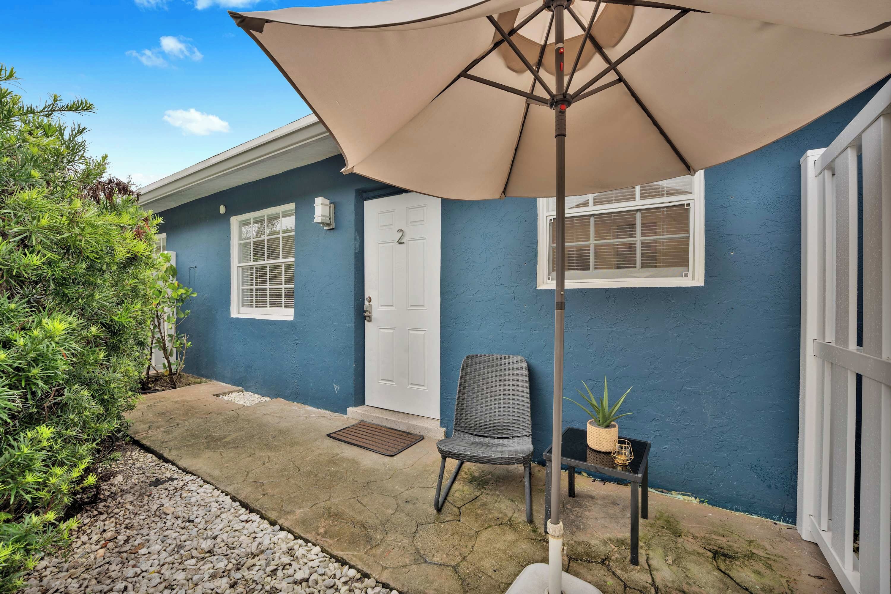 A house with a blue exterior, a white door, and a window with blinds. There is a chair and a small table with a plant.
