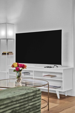 En moderne stue med et fladskærms-tv på et hvidt tv-møbel, et sofabord i glas og en vase med blomster.