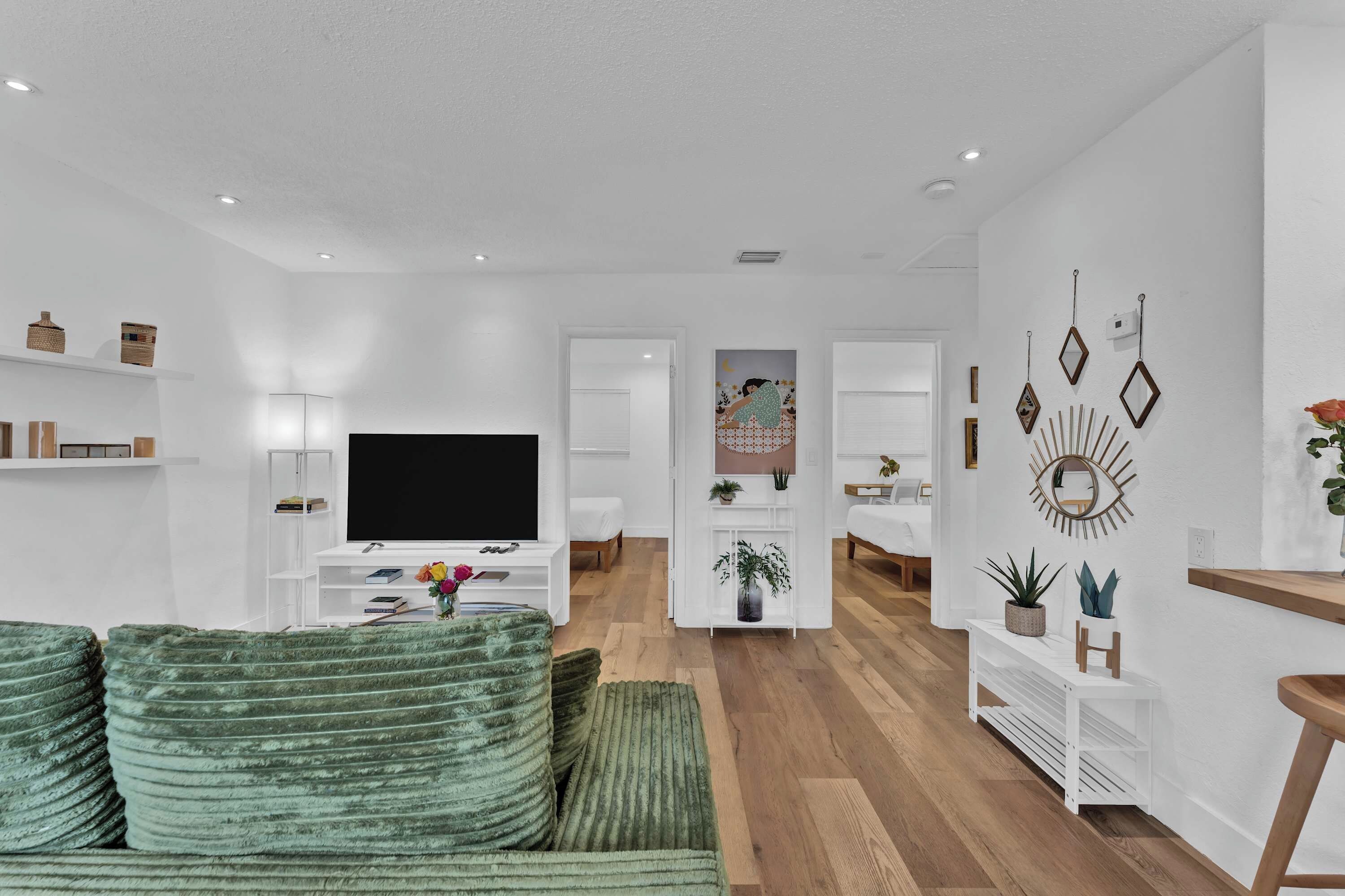 A modern living room with a flat-screen TV, green sofa, white shelves, and a hallway leading to a bedroom.