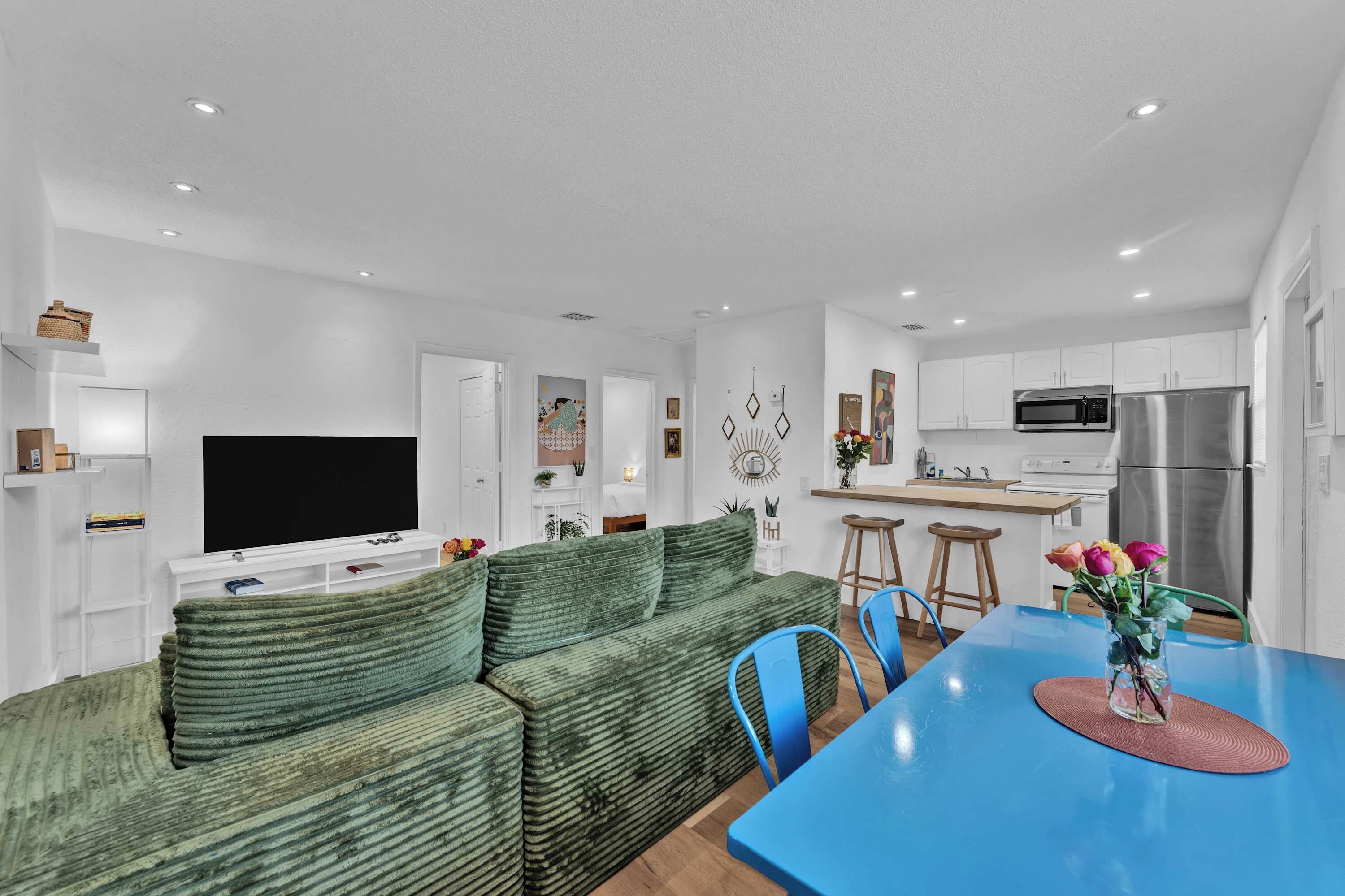 A modern living room with a green sofa, a flat-screen TV, and a blue dining table with chairs.