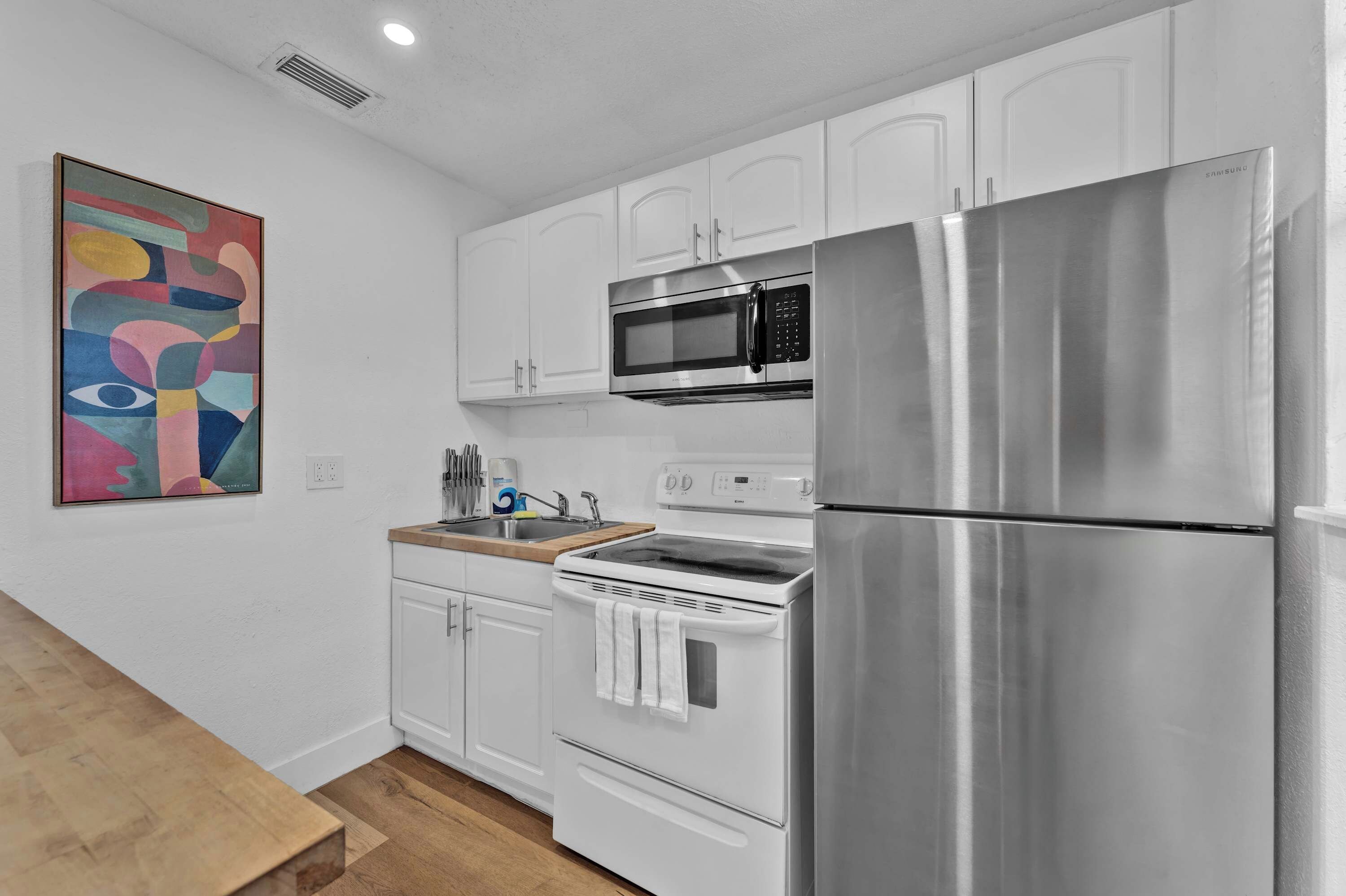 A compact kitchen with white cabinetry, a stainless steel refrigerator, a microwave, an oven, and a sink.