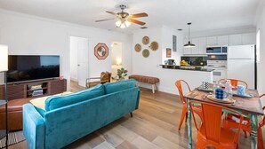 A modern living room with a teal sofa, a dining table with orange chairs, a kitchen area with white cabinets, and a ceiling fan. - Sunny Side Stay Condo- 2 bed 1 bath Near Riverwalk (Fort Lauderdale)