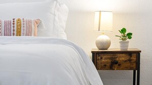 A neatly made bed with a white comforter and a decorative pillow. A bedside table with a lamp and a potted plant is visible. - Sunny Side Stay Condo- 2 bed 1 bath Near Riverwalk (Fort Lauderdale)