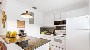 A modern kitchen with white cabinets, a granite countertop, a white refrigerator, and a hanging pendant light. - Sunny Side Stay Condo- 2 bed 1 bath Near Riverwalk (Fort Lauderdale)