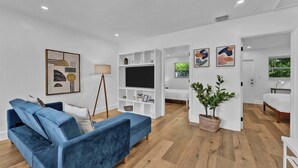 A modern living room with a blue sofa, a flat-screen TV, a bookshelf, and a potted plant. - Flamingo Escape: Bright 2BR Near Riverside Beach (Fort Lauderdale)