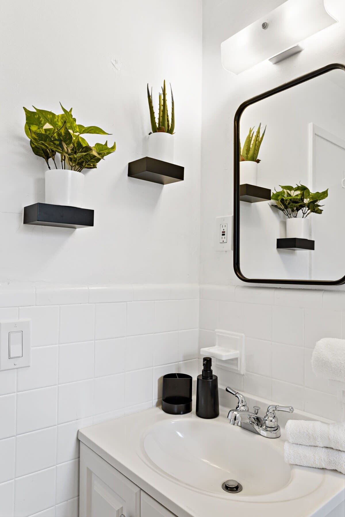 Un bagno moderno con piastrelle bianche, un lavandino bianco, uno specchio e due mensole nere sospese con piante.