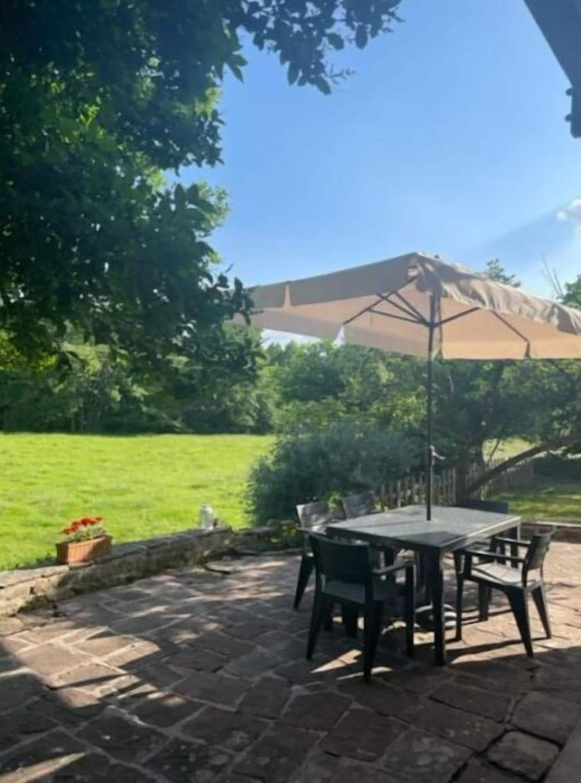Une terrasse extérieure avec une table et des chaises, une pergola offrant de l’ombre et une vue sur une pelouse verdoyante et des arbres.