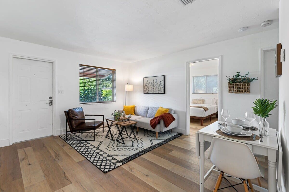 Ein modernes Wohnzimmer mit einer Couch, einem Esstisch und einem Couchtisch. Es gibt ein Fenster mit Jalousien, eine Tür und ein Wandregal mit Pflanzen.