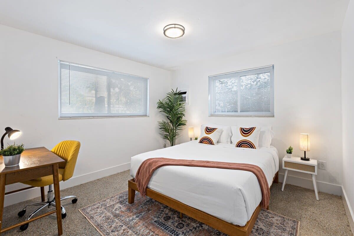 A bedroom with a bed, a desk, a chair, a lamp, and a plant.