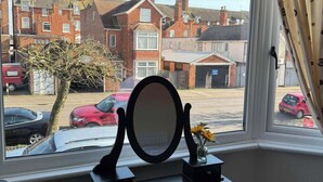 A bedroom with a window view of a residential street, a bedside table with a mirror, and a small vase with flowers. - The Singlecote Hotel (Skegness)