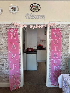 Uma cozinha com parede de tijolos, uma placa de boas-vindas e dois banners com os dizeres 'Feliz Dia das Mães' e 'DIA DAS MÃES'.