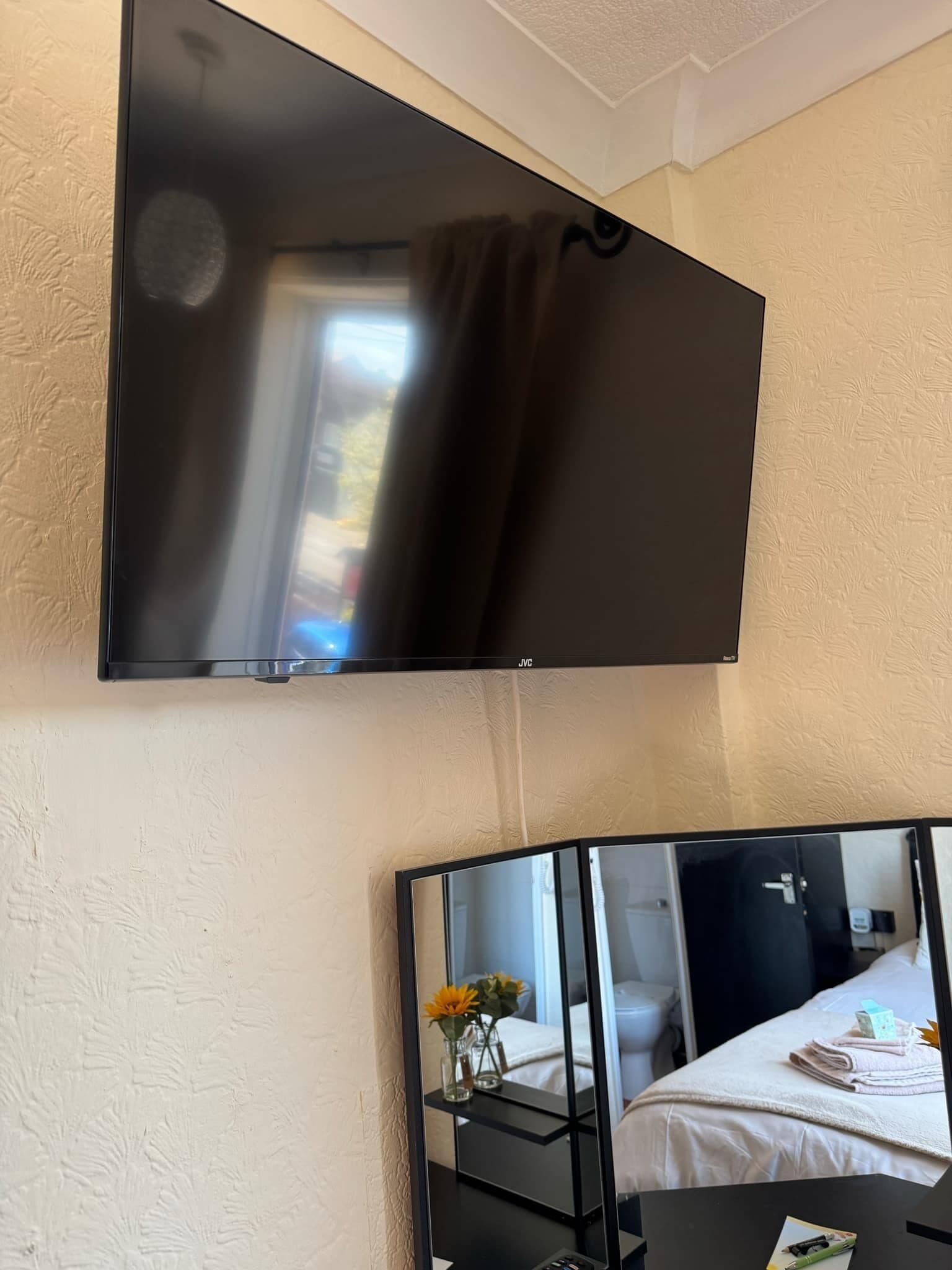 Una habitación con una televisión de pantalla plana montada en la pared, una cama con edredón blanco y una mesita de noche con un jarrón de flores.