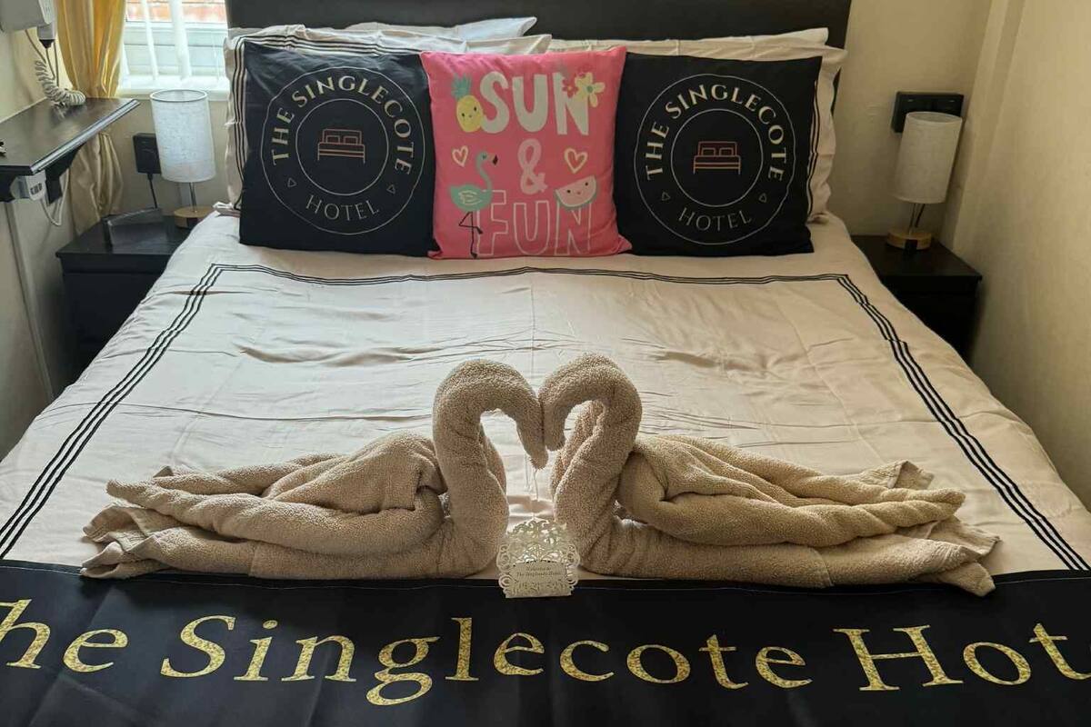 A hotel room with a bed featuring a towel art of two hearts, pillows with the hotel logo, and a towel rack with a towel.