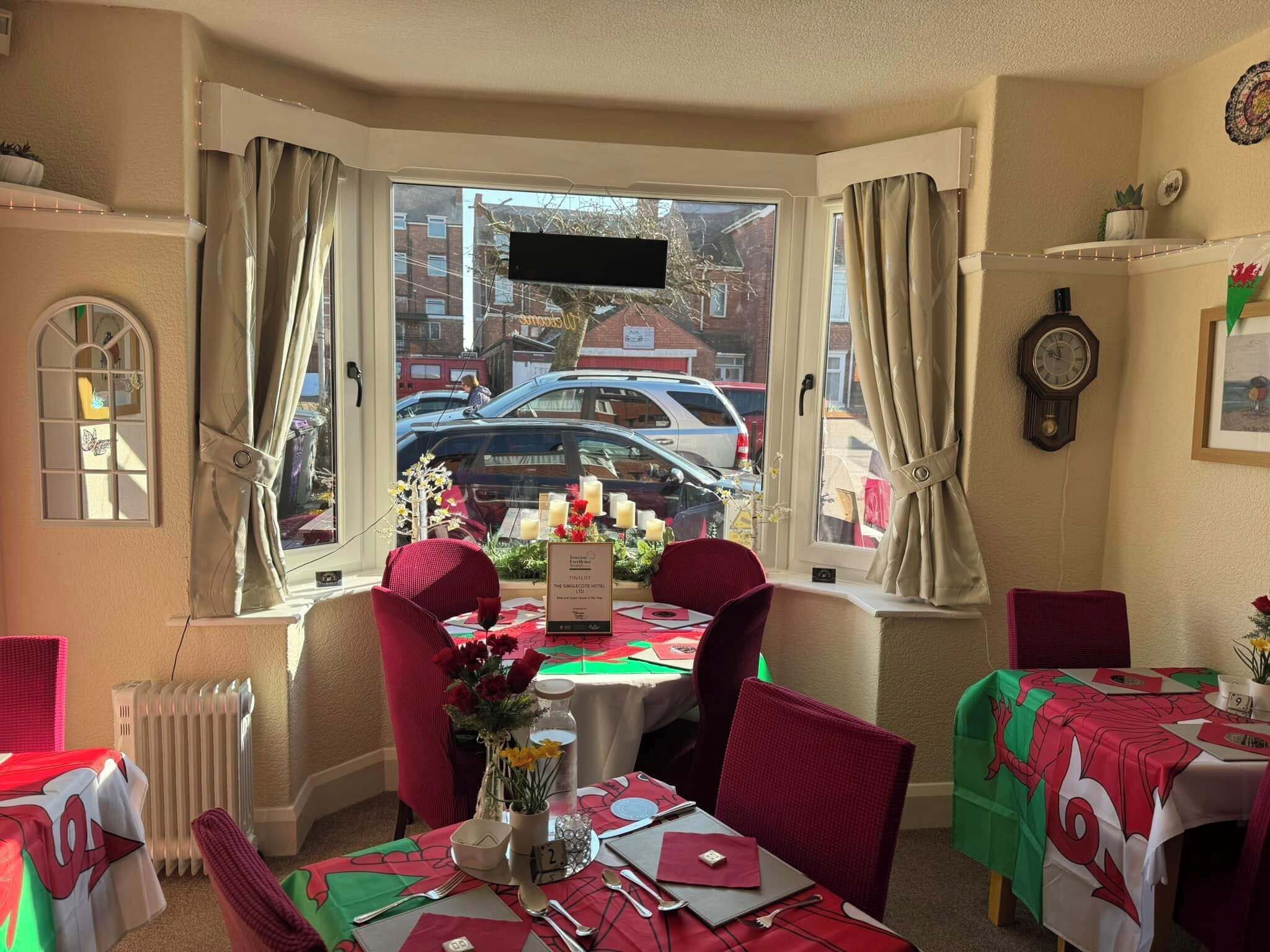 Un coin repas avec une table dressée pour deux personnes, une fenêtre avec des rideaux, un radiateur et une horloge au mur.