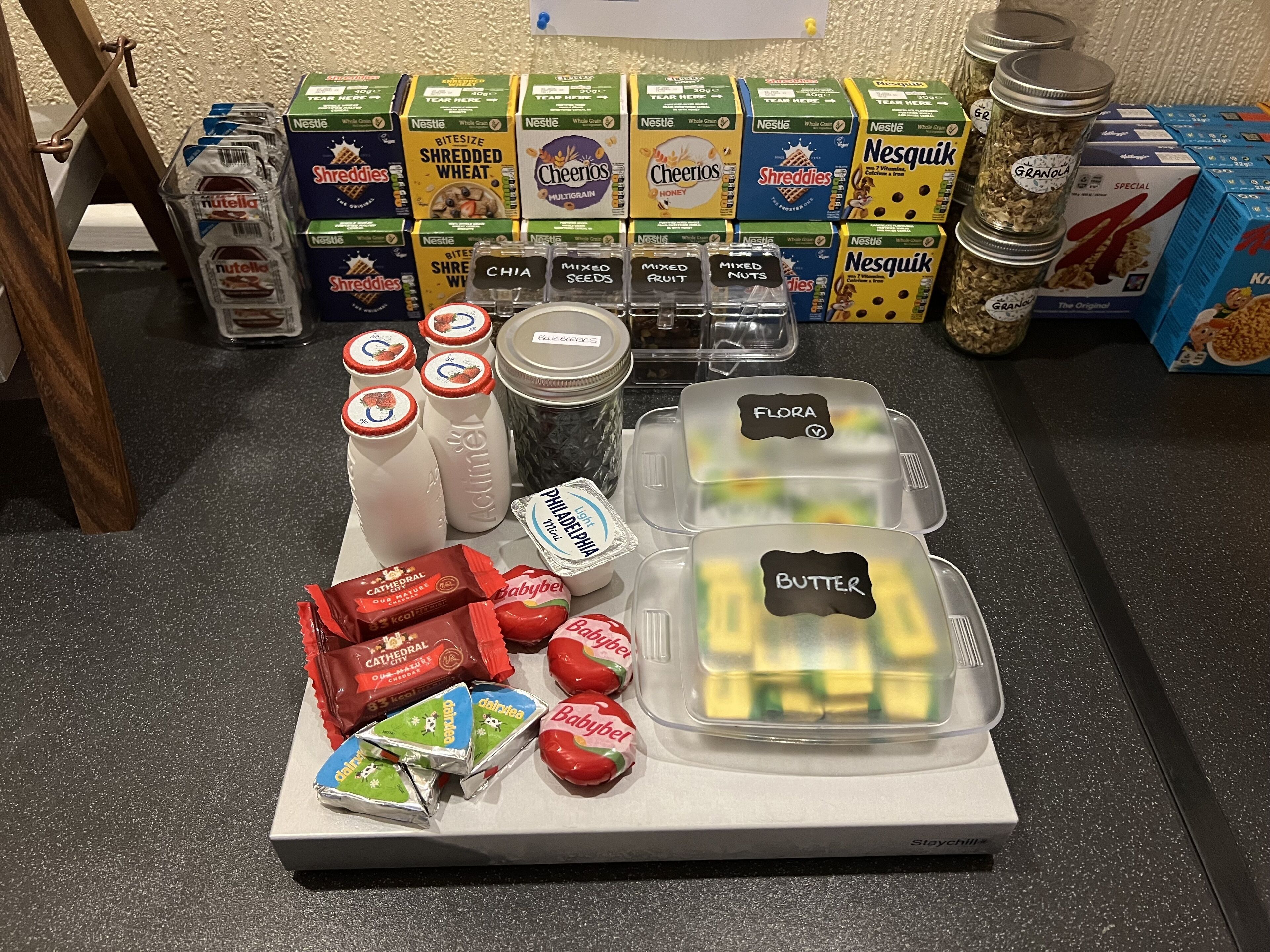Una encimera de cocina con diversos alimentos y recipientes, incluyendo cajas de cereales, frascos de frutos secos y envases con ingredientes como 'Cheerios' y 'Butter'.