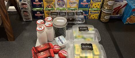 Una encimera de cocina con diversos alimentos y recipientes, incluyendo cajas de cereales, frascos de frutos secos y envases con ingredientes como 'Cheerios' y 'Butter'.