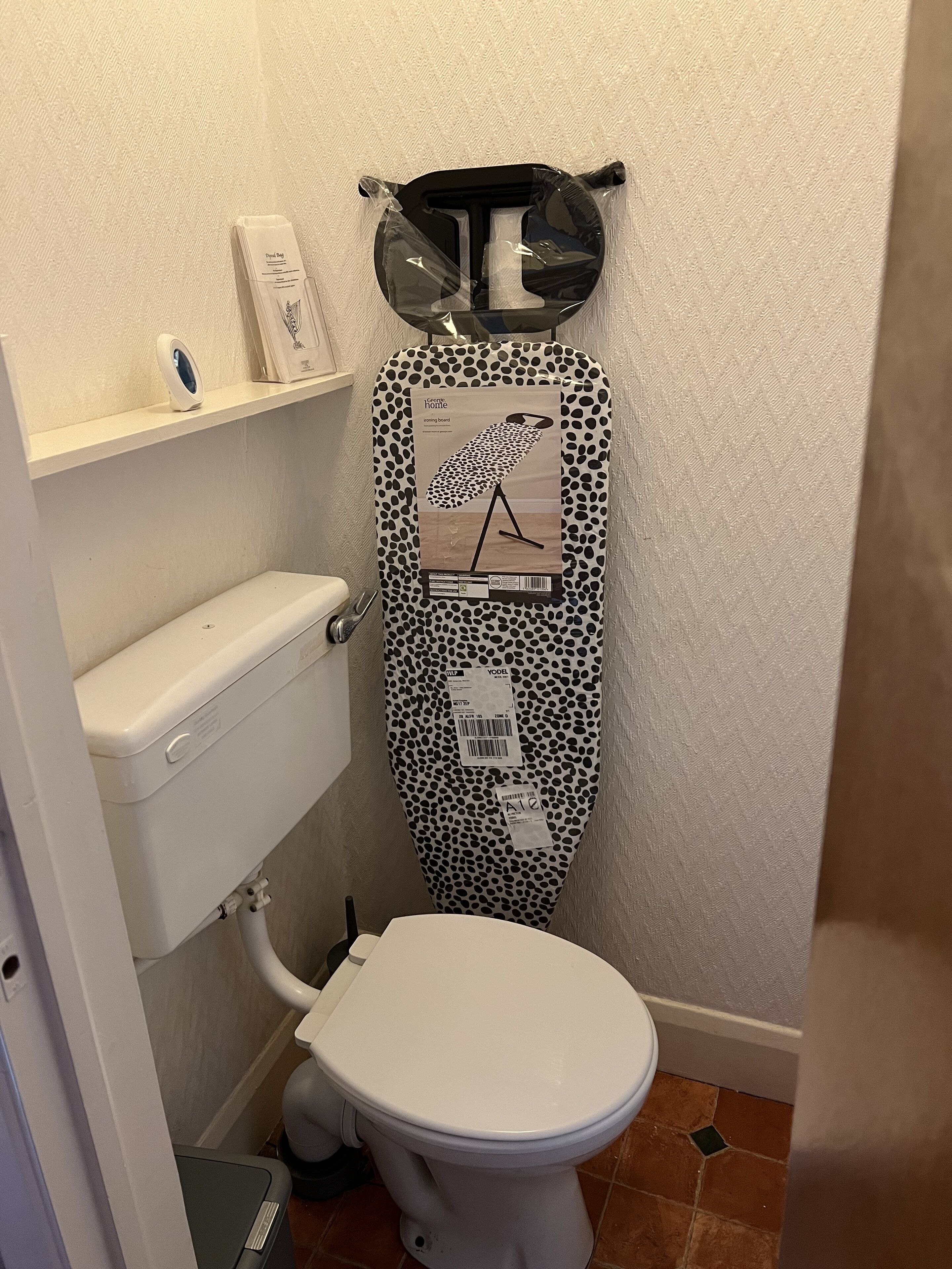 A bathroom with a toilet, a white shelf with a small device, and a polka-dotted ironing board.