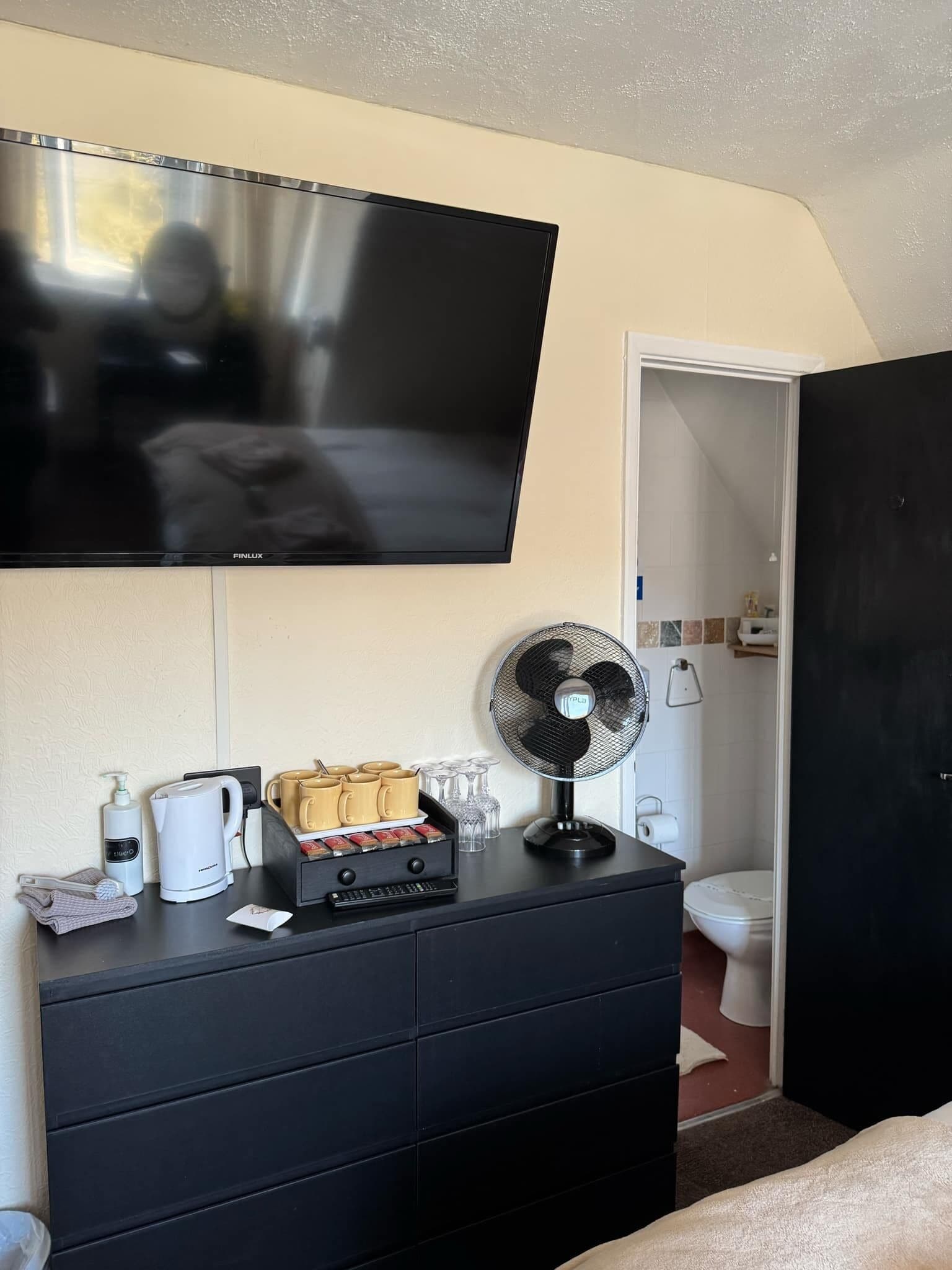 A hotel room with a flat-screen TV mounted on the wall, a black dresser with a coffee maker, a kettle, and a tray of coffee cups. There is a standing fan on the dresser and a bathroom visible through an open door.