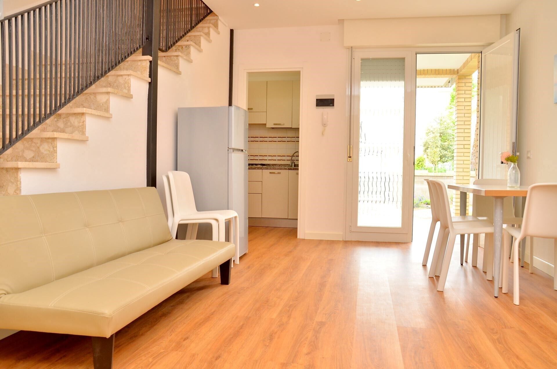 Una sala de estar moderna con sofá, sillas, una escalera, cocina y un ventanal corredero que da a un área exterior.
