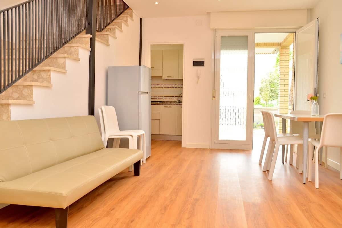 Ein modernes Wohnzimmer mit Sofa, Sesseln, einer Treppe, einer Küche und einer Schiebeglastür zu einem Außenbereich.