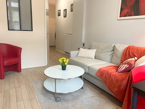 Una sala de estar moderna con una butaca roja, un sofá blanco, una mesa redonda de centro con flores amarillas y una alfombra gris.