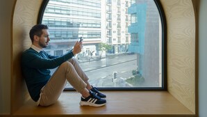 Une personne assise près d’une grande fenêtre en arc d’où l’on aperçoit un paysage urbain.