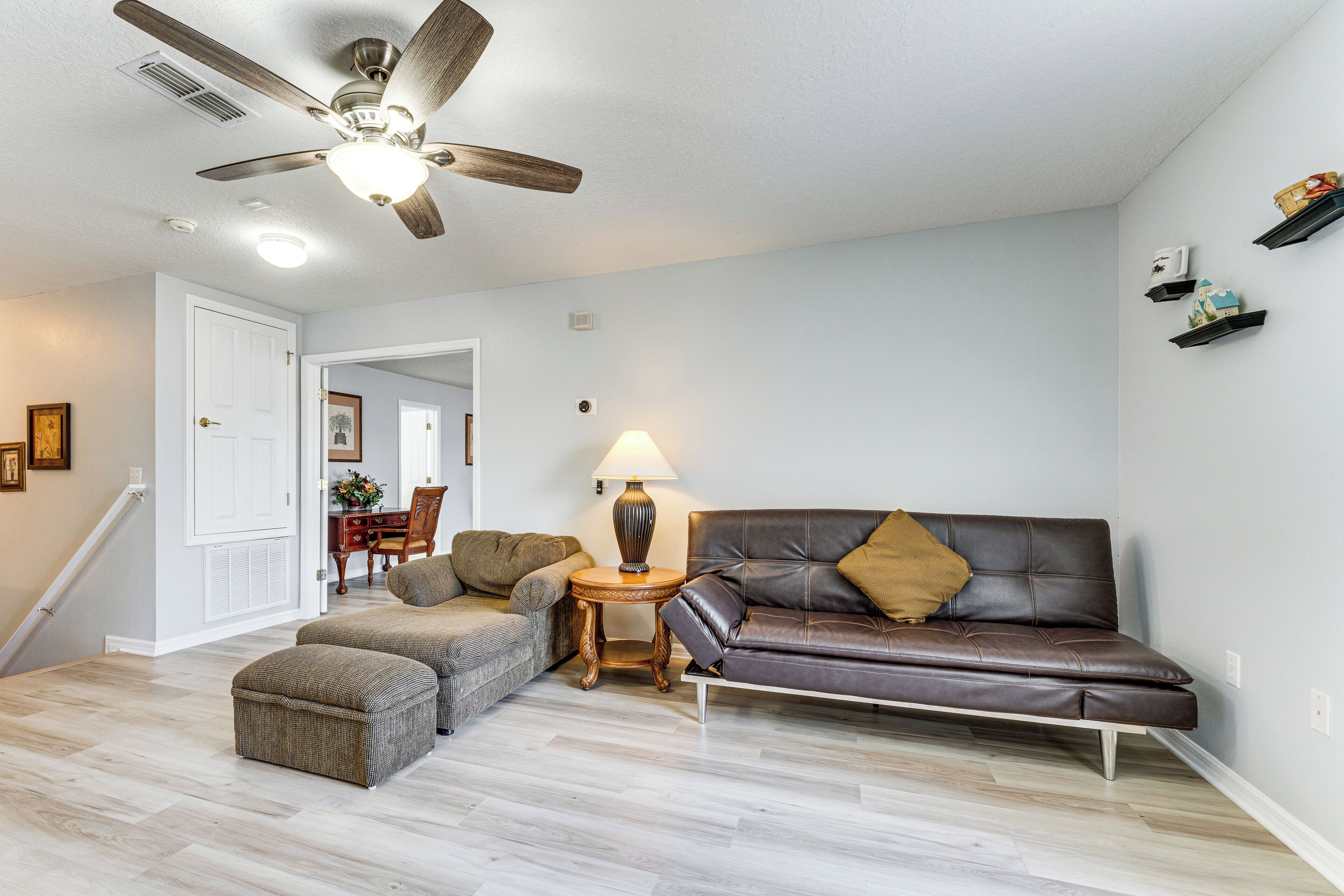 Un salon avec un ventilateur de plafond, un canapé en cuir marron, un fauteuil à motifs, une petite table d’appoint et une lampe.